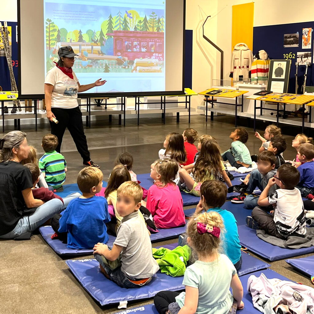 Atelier scolaire présenté sur un écant géant devant des enfants assis au sol sur des tapis bleus.