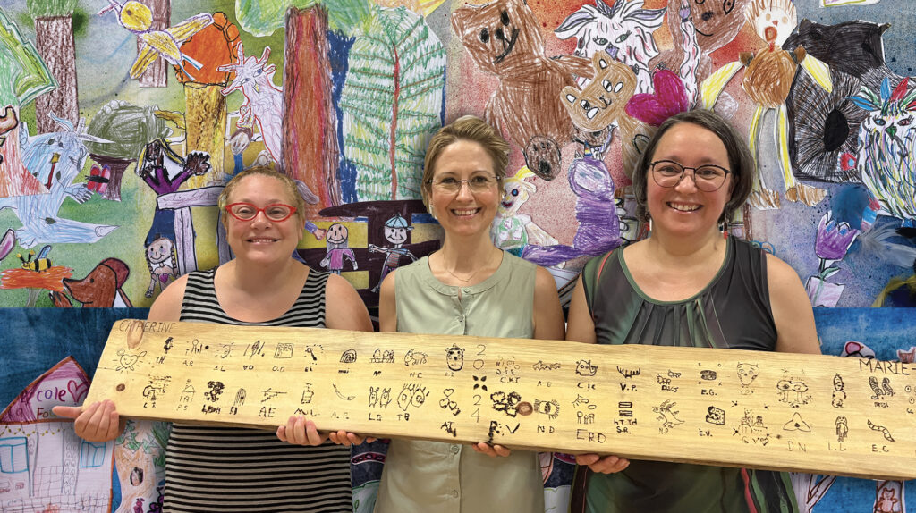 Isabelle Charbonneau accompagnée de deux enseignantes qui présente une planche couverte d'icones pyrogravés par des enfants devant une murale de dessins d'enfants.