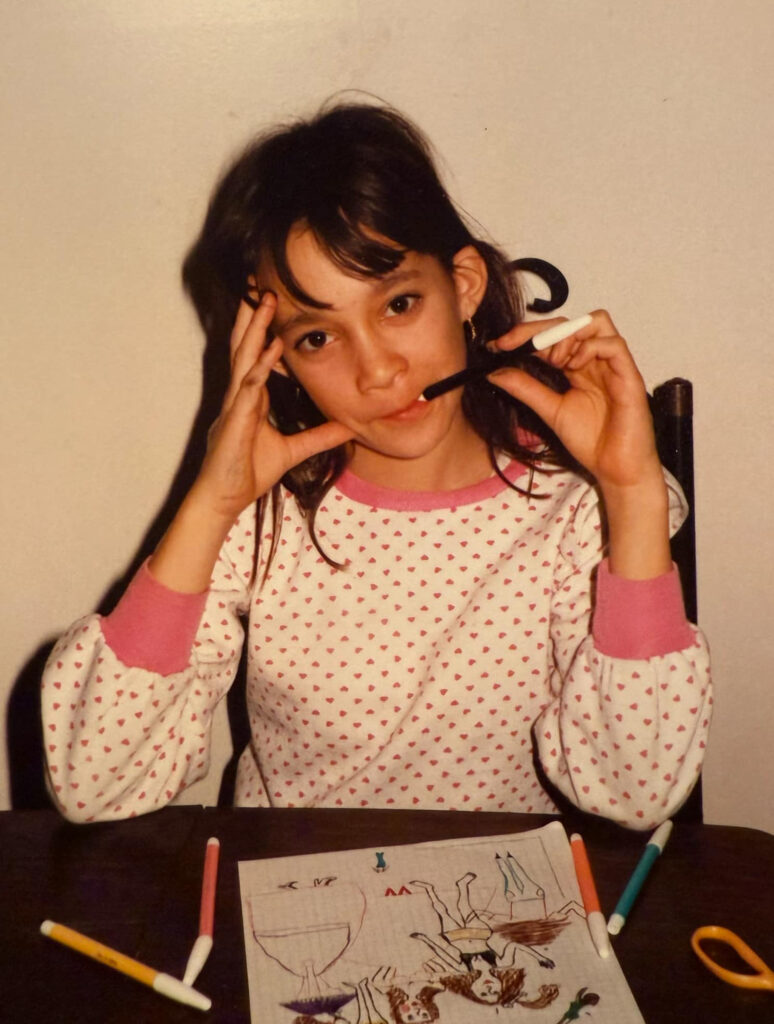 Isabelle Charbonneau à 9 ans avec un crayon dans la bouche, assise devant une table où l'on peut voir un coloriage.